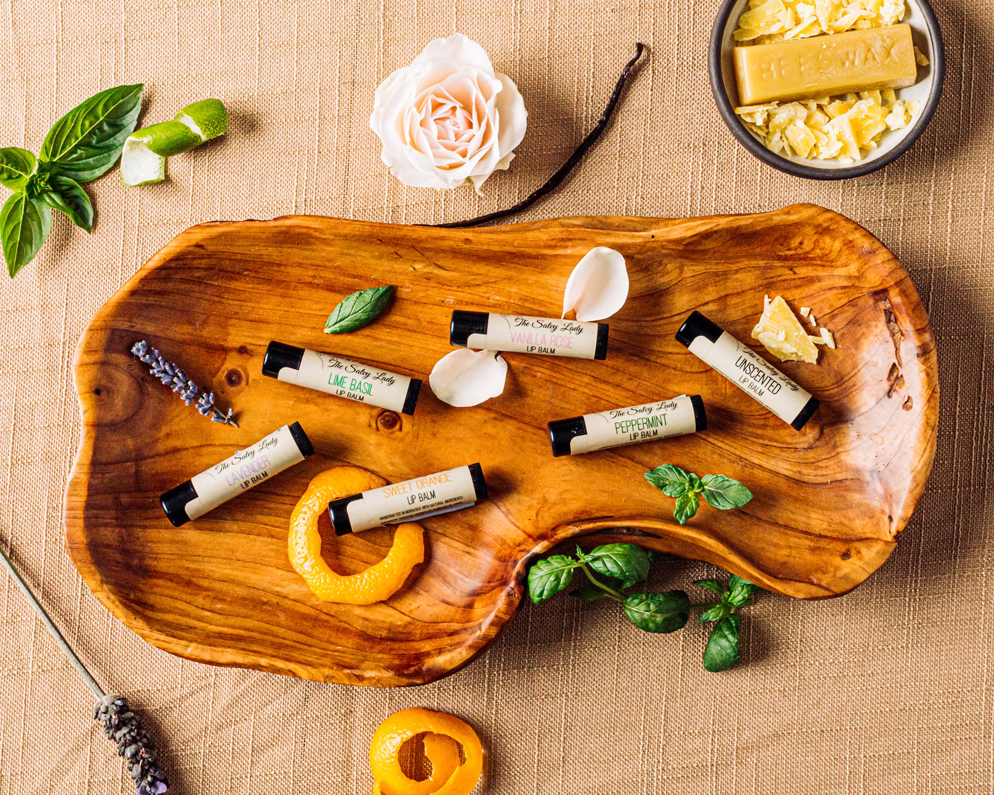 A wooden bowl set on a burlap cloth contains 6 lip balms. The natural ingredients that were used to make the balms are scattered throughout the photo.